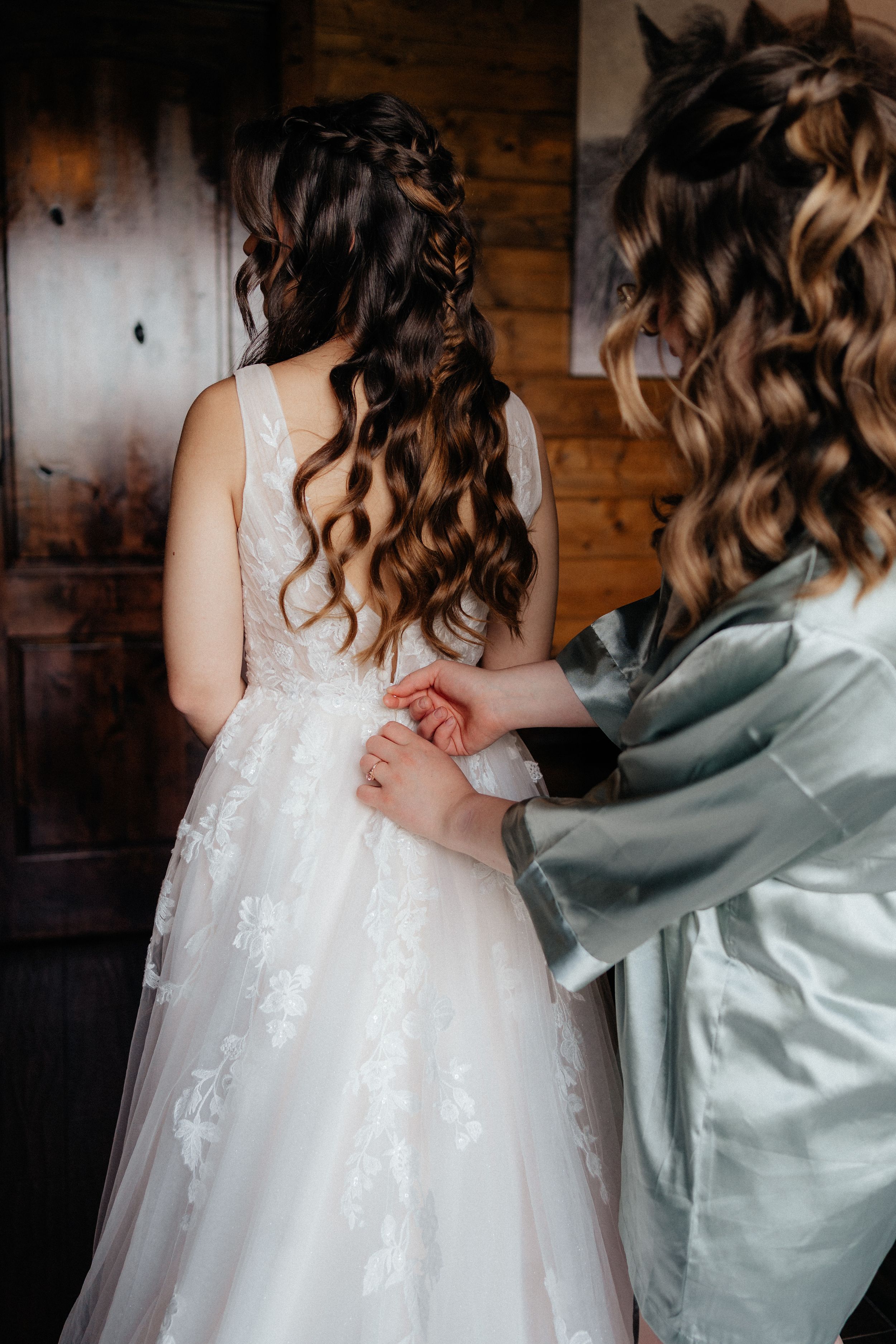 Bridesmaid in sage robe fastens the back of bride's lace wedding gown in a rustic wooden room.