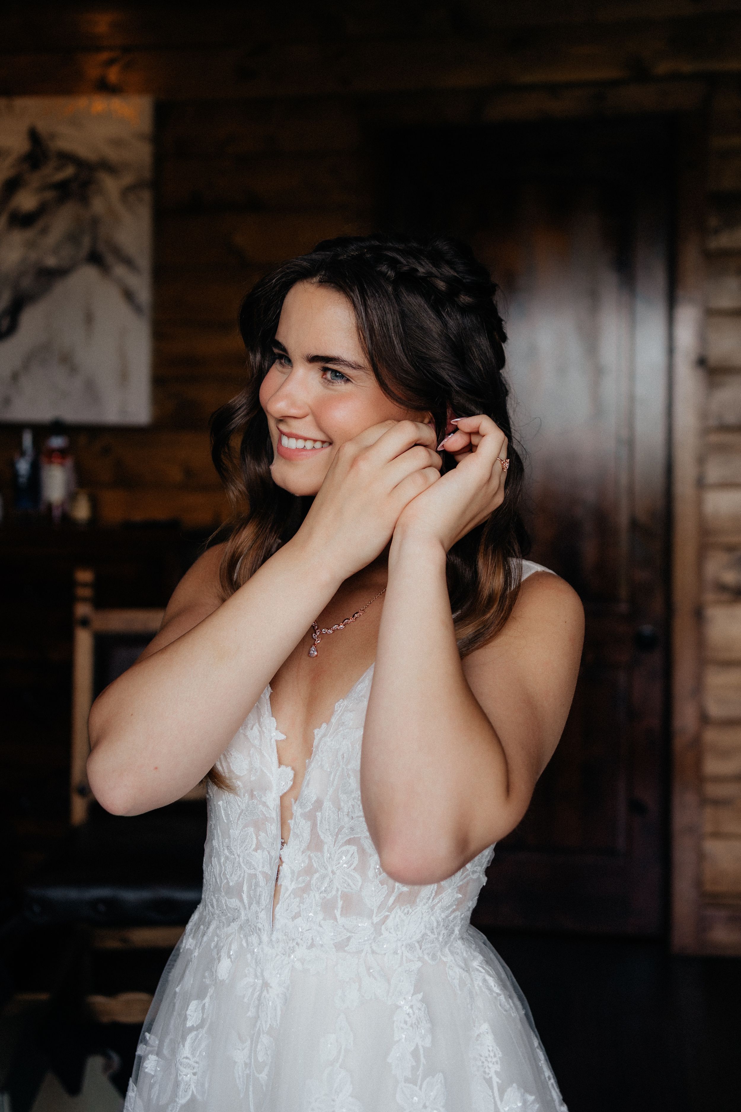 Smiling bride in lace gown puts on earrings in rustic wooden interior.