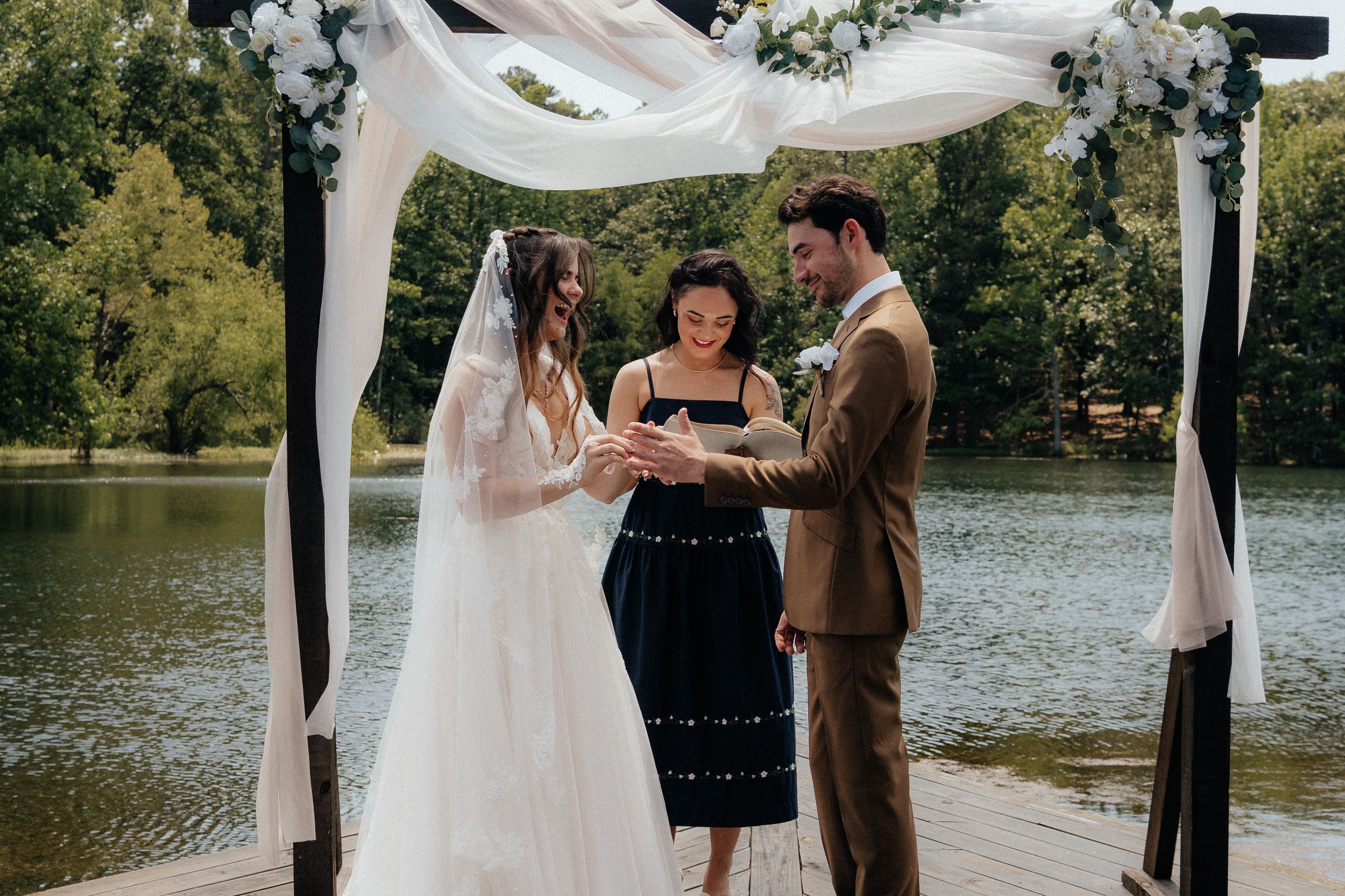 Bride and groom exchanging rings at outdoor lakeside wedding ceremony under floral arch with officiant.