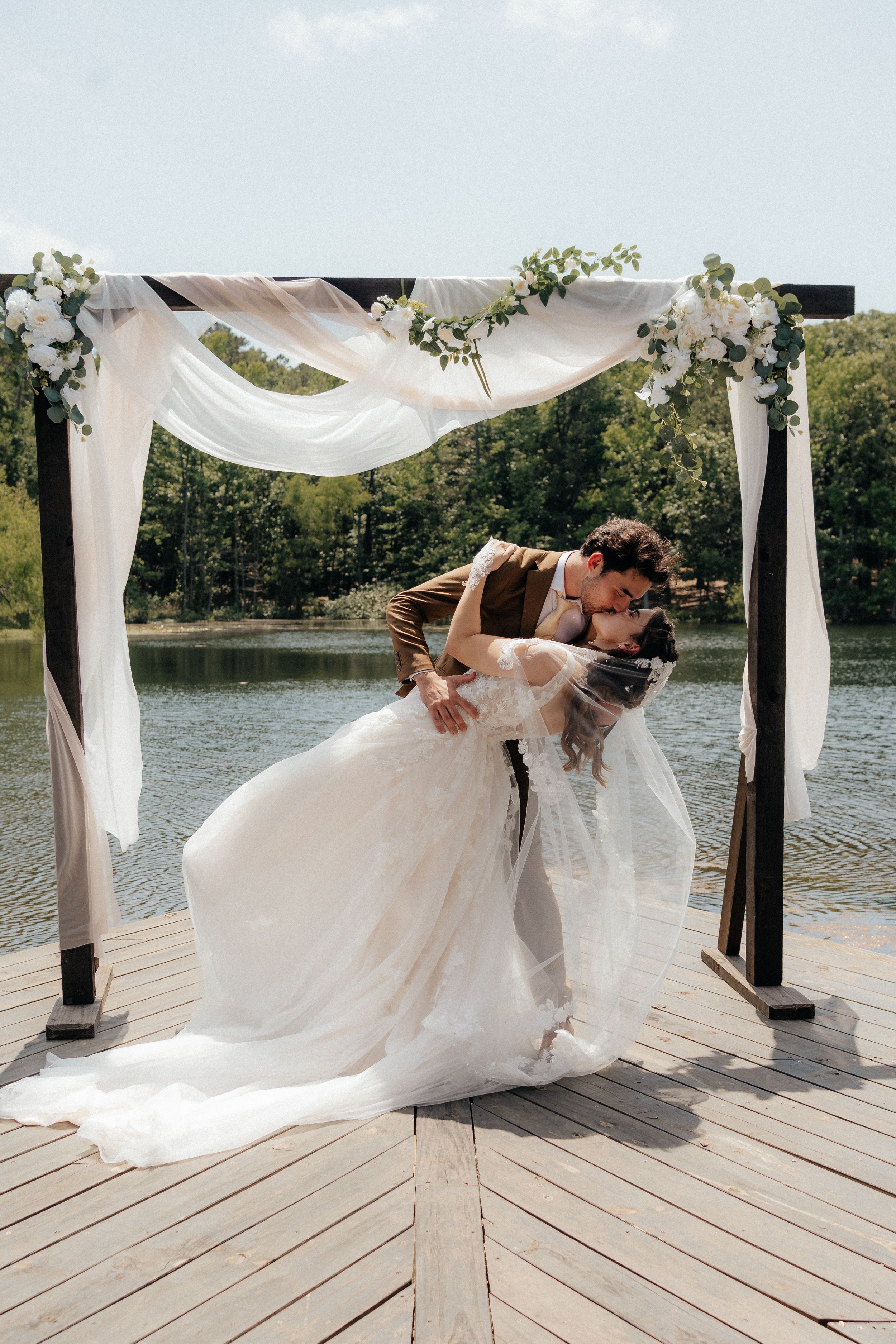 Bride and groom sharing a kiss under a floral arch on a lakeside wooden dock on their wedding day.