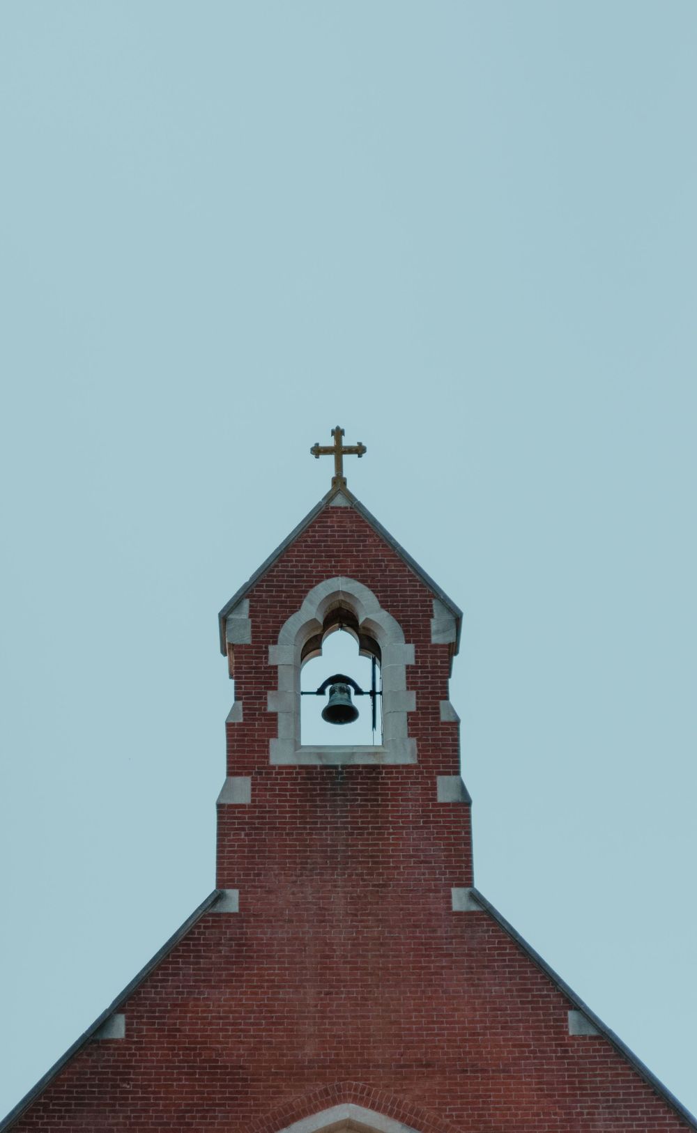Red brick church bell tower with cross against light blue sky.