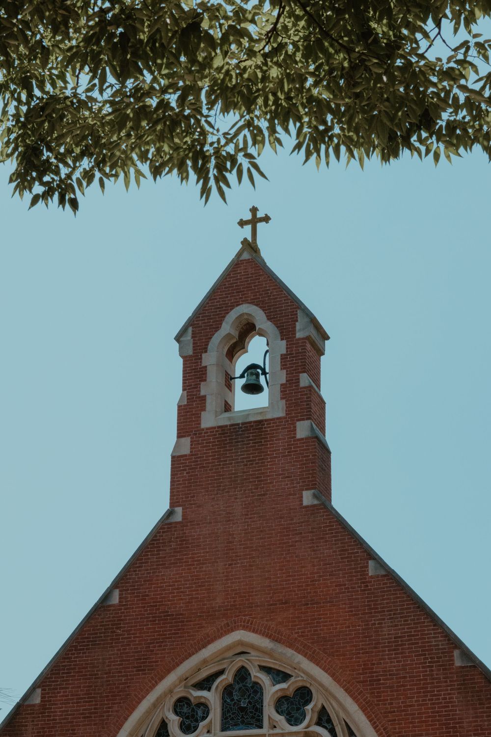 Gothic church steeple with bell tower framed by green leaves against blue sky.