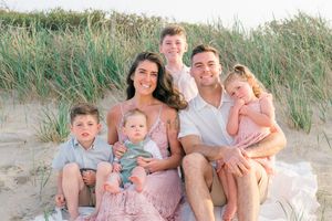 Relaxed family portrait on beach at sunset, tall grass and warm glow creating dreamy setting.