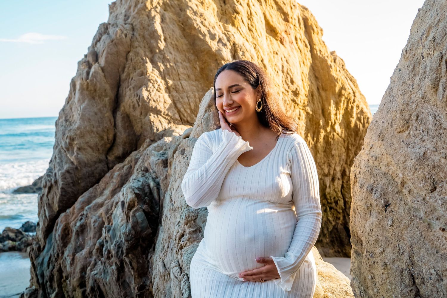 A maternity portrait of an expectant mother in white dress posing against dramatic coastal rock formations. malibu.