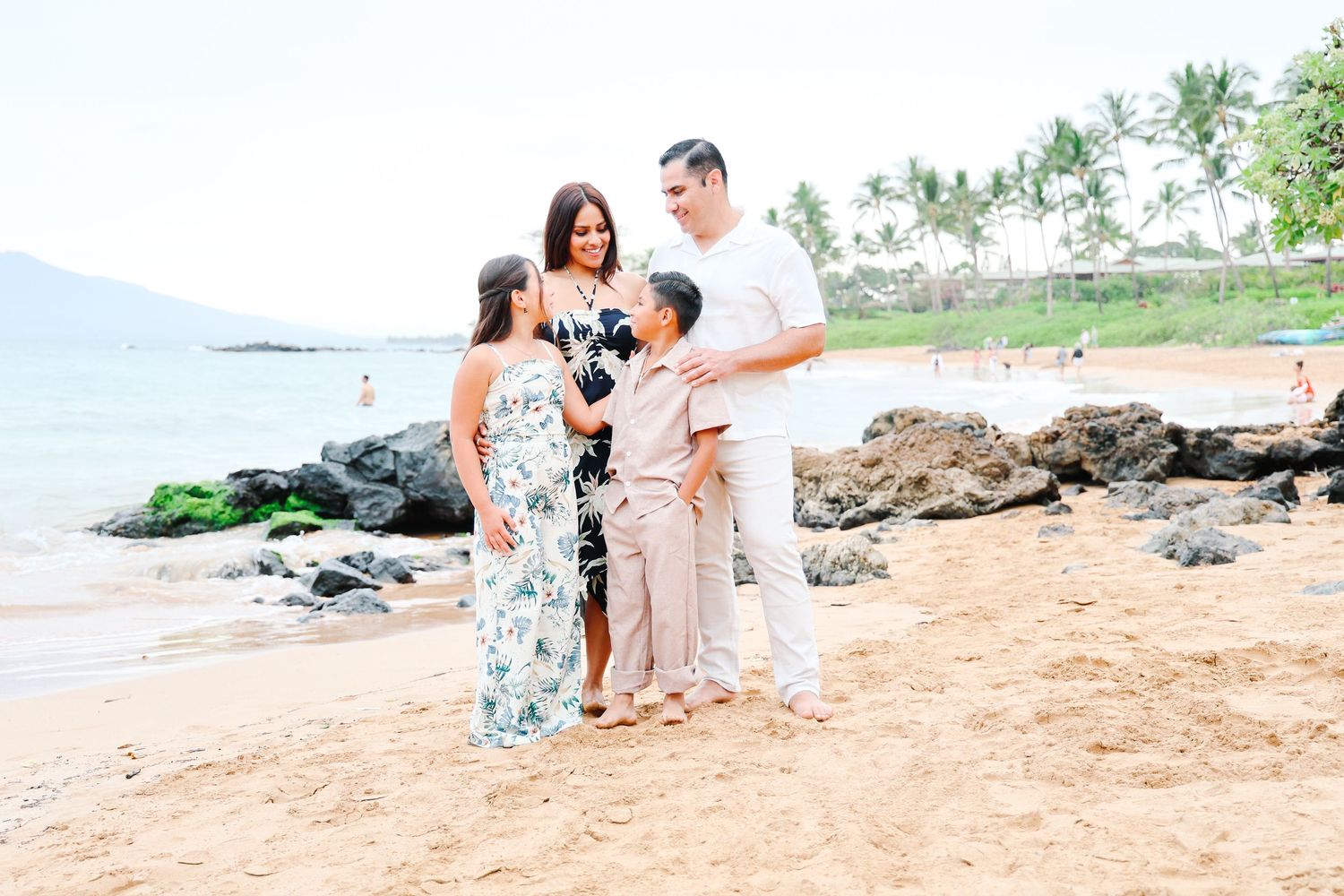 A couple in white and floral outfits holds hands while walking on a sunny beach.
