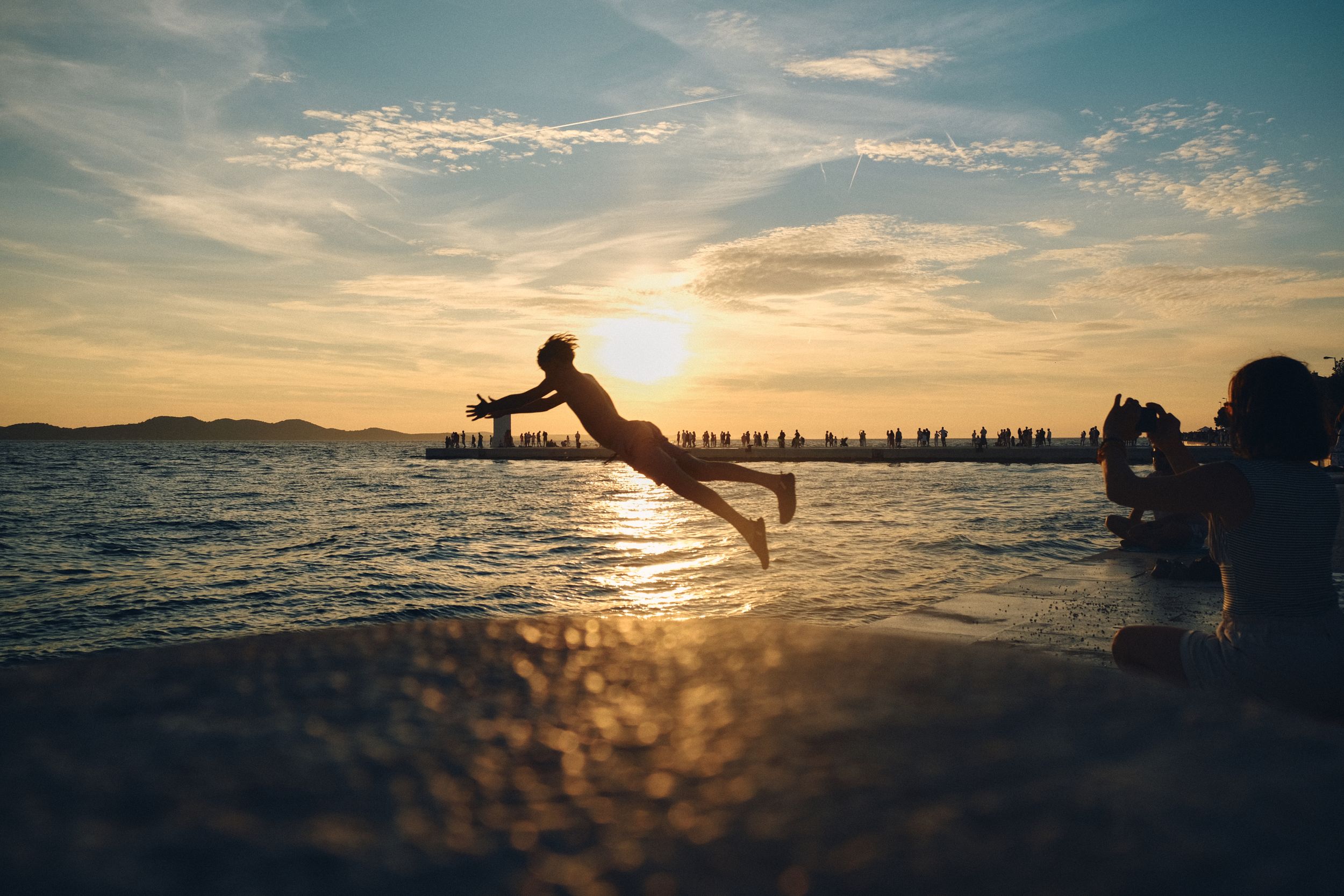 Silhouette of person jumping into ocean at sunset with city skyline in background.