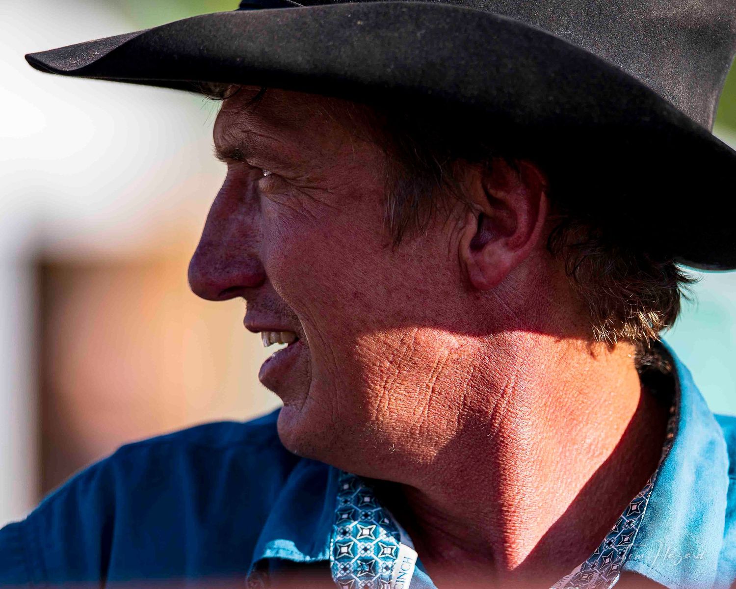 A weathered cowboy wearing a black hat in profile, with sun-kissed skin and a blue western shirt.