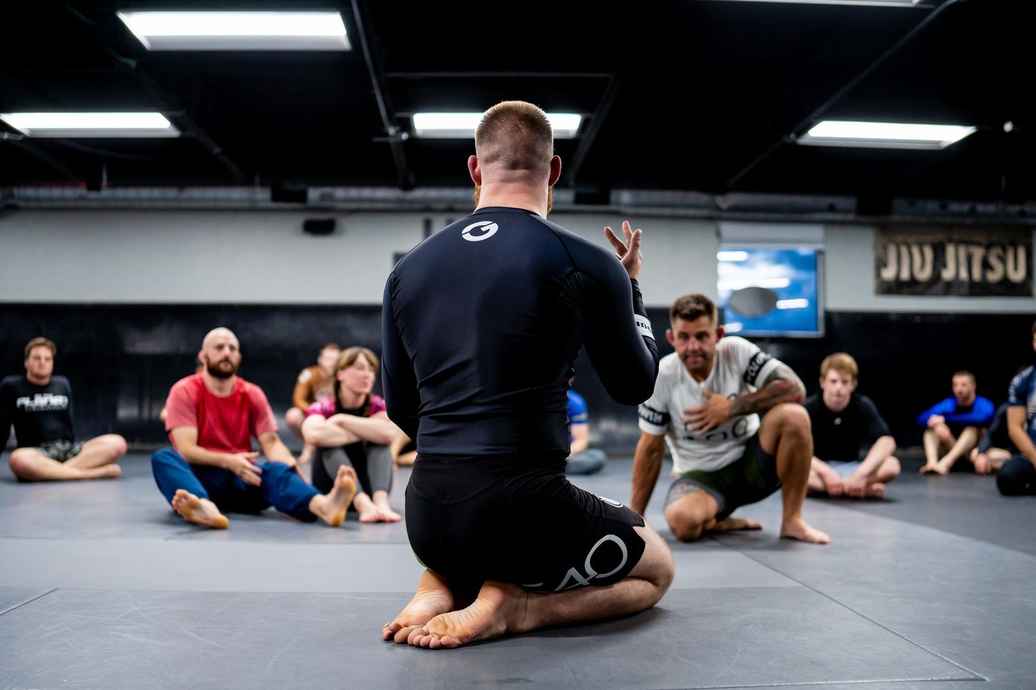 Students sit on a mat while attending a martial arts instruction class in a gym.