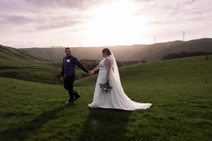 Groom looking back at the bride holding hands while walking in open field and backlit from the sun against rolling hills