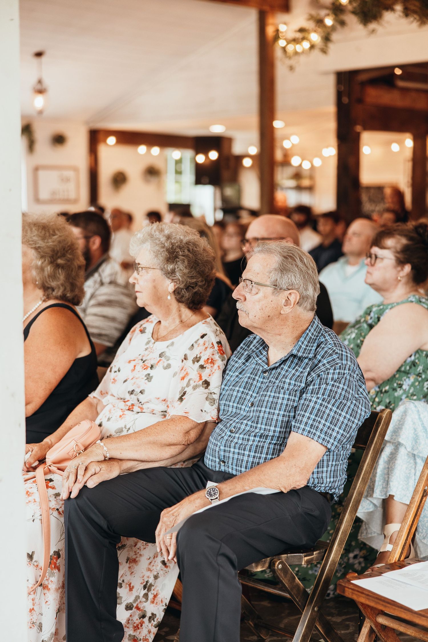 Grandparents holding hands during their granddaughter’s wedding ceremony. Beautiful floral dress and beautiful lighting in the background.