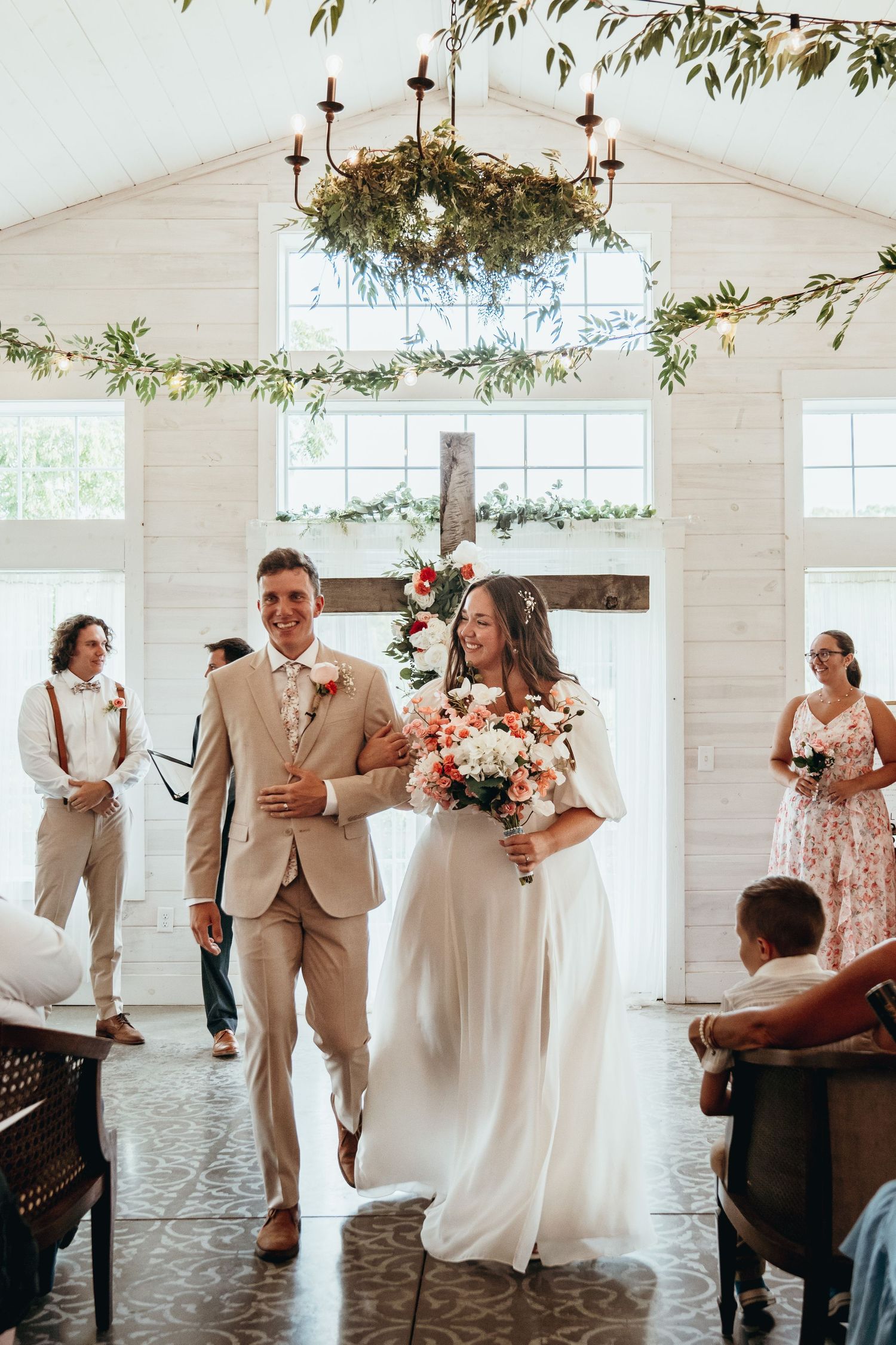 Beige Groom Suit. Pink Wedding Bouquet. Bride and Groom exiting wedding ceremony after their First Kiss. Beautiful white and green venue.
