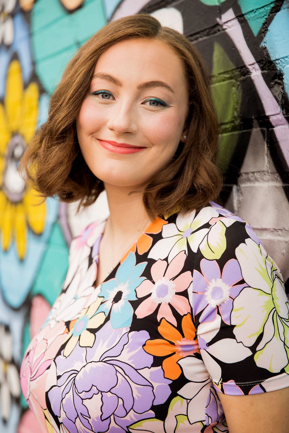 Young woman in colorful floral dress smiles in front of vibrant graffiti wall mural.