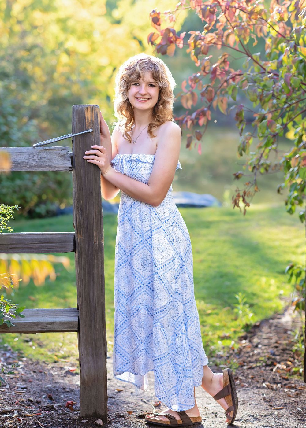 Young girl in blue and white maxi dress poses by wooden fence in autumn garden.