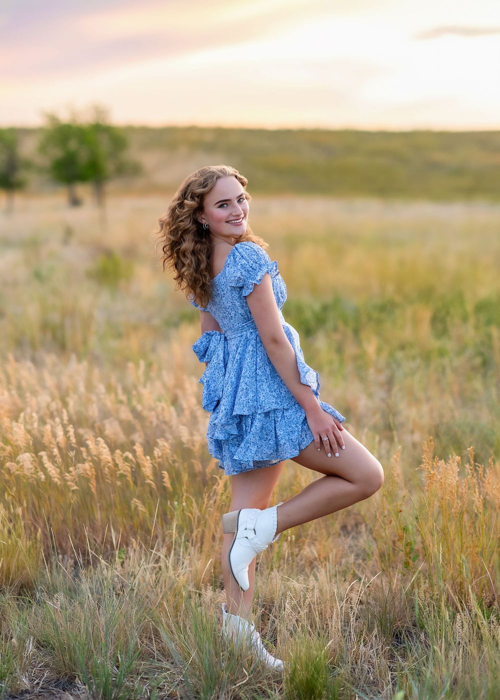 Curly-haired girl in blue ruffled dress and white boots poses in golden field at sunset.
