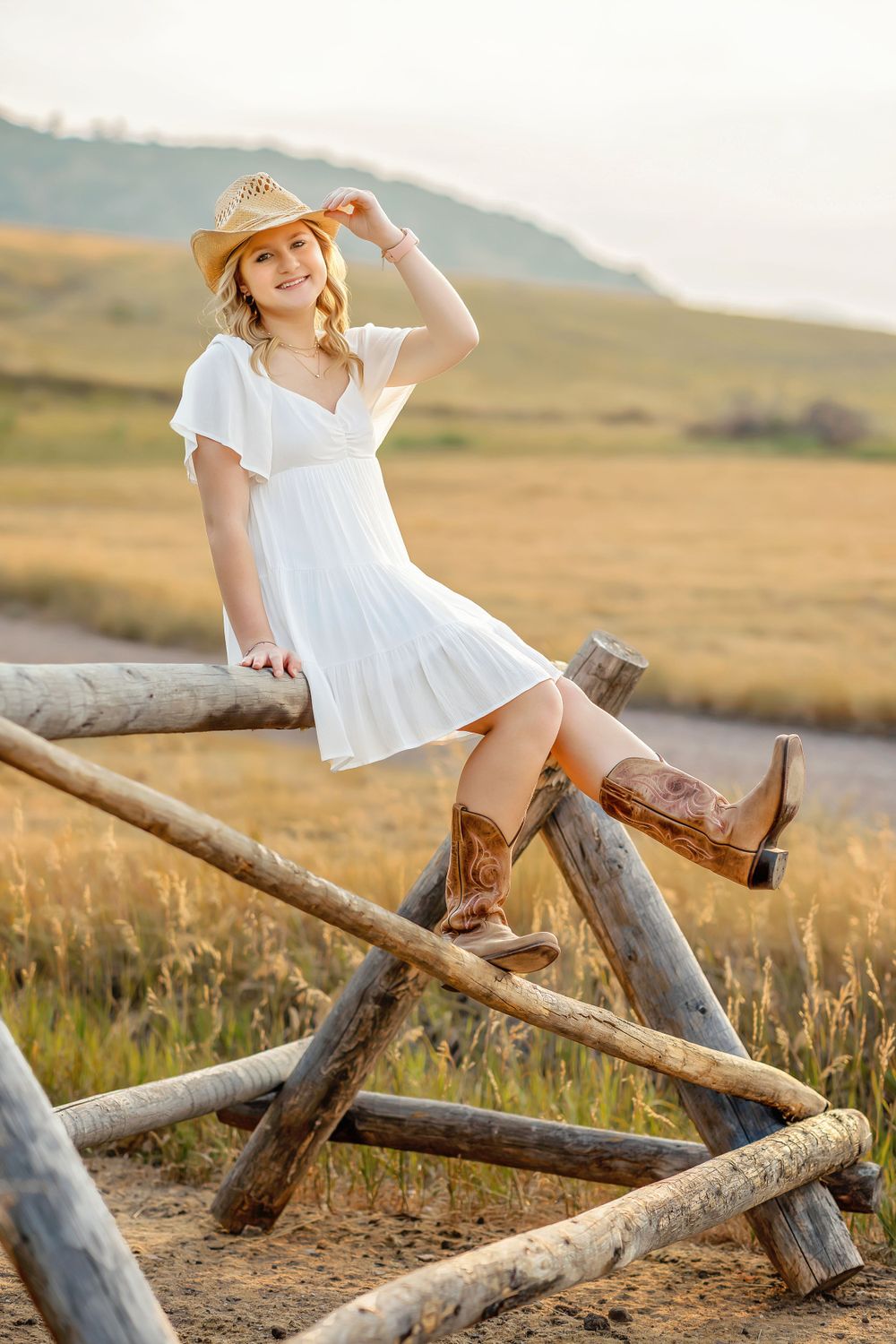 Young woman in white dress and cowboy hat sitting on wooden fence in golden field.