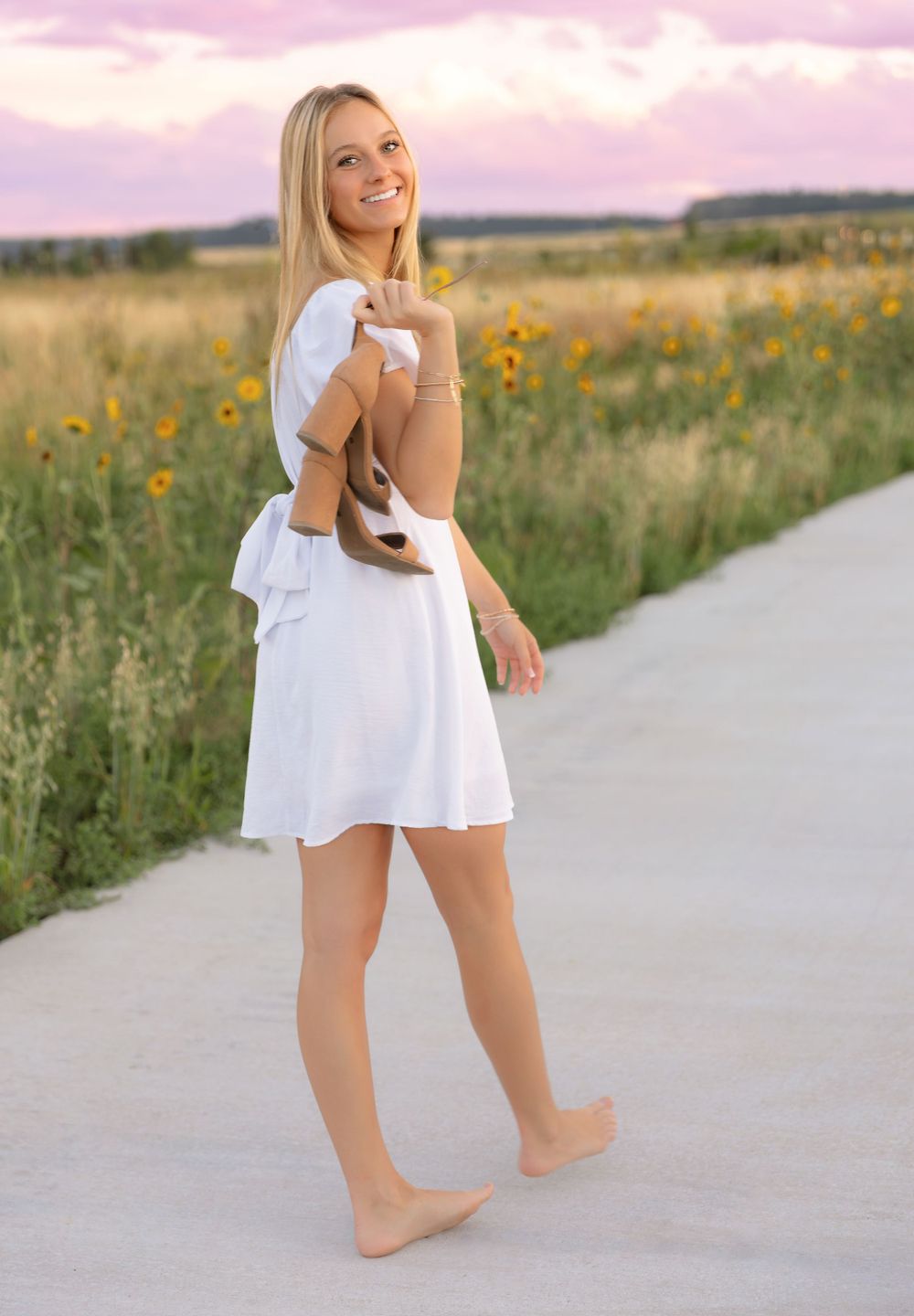 Young blonde woman in white dress walking barefoot on a path surrounded by sunflowers at sunset.
