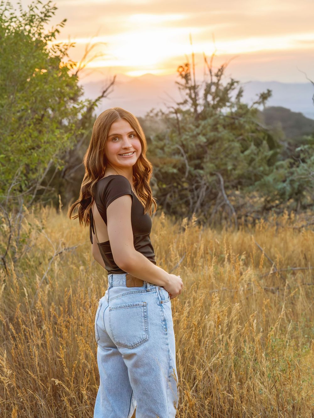 Smiling teenage girl in black crop top and jeans poses in golden field at sunset.