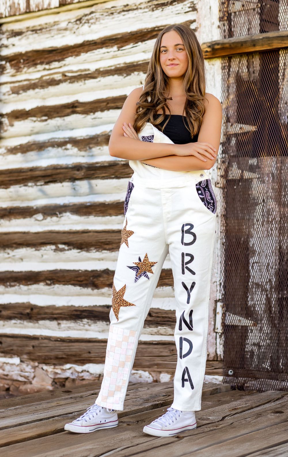 Teen girl in custom white overalls reading Brynda posing by rustic log cabin wall.