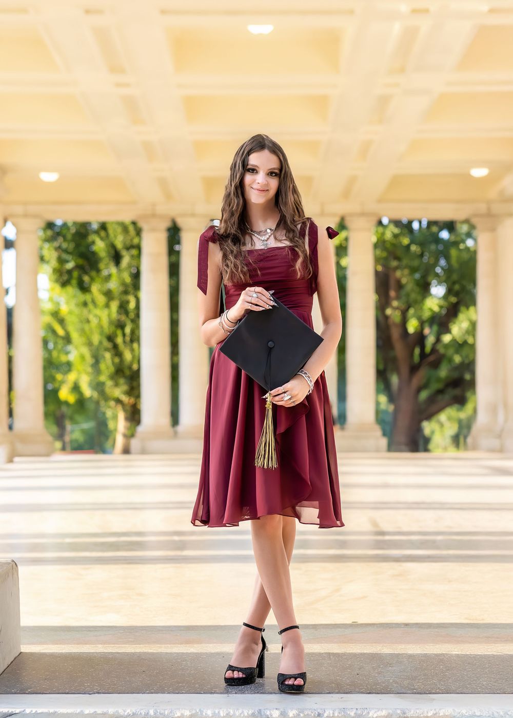 Young woman in burgundy dress holds graduation cap while posing between elegant columns.