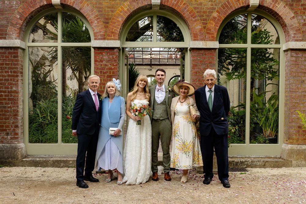 A wedding party poses for photos outside a historic brick building with arched windows and lush greenery.