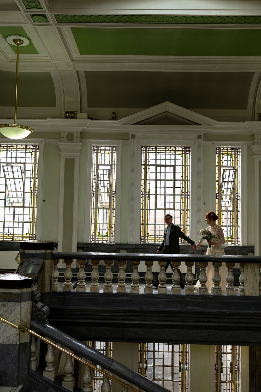 Two people walk along an ornate interior balcony with stained glass windows and classical architectural details.