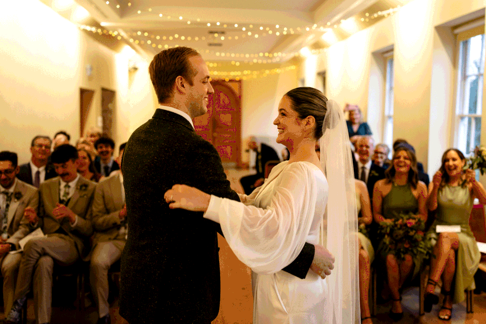 A couple shares their first dance at an indoor wedding reception as guests watch from their seats along the walls.