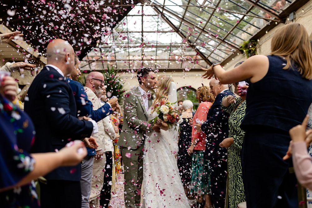 Wedding guests celebrate under a greenhouse roof as pink flower petals rain down during a joyful reception moment.