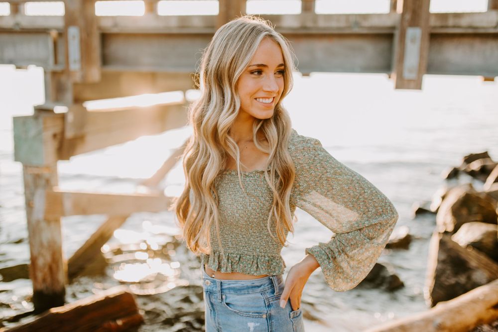 Person wearing sparkly grey top and jeans poses by waterfront at golden hour.