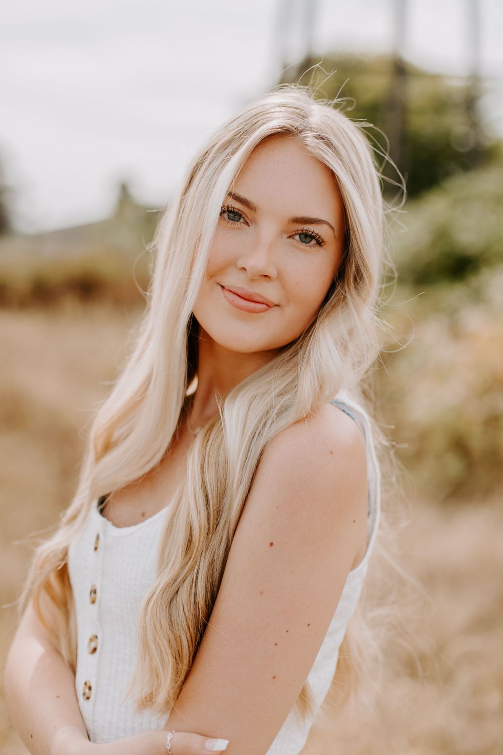 Portrait of a person with long blonde hair in a white button-up top against a natural outdoor background.