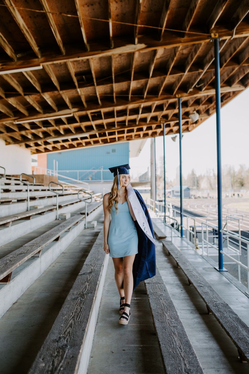 Graduate in blue dress and cap walking under covered bleachers on graduation day.