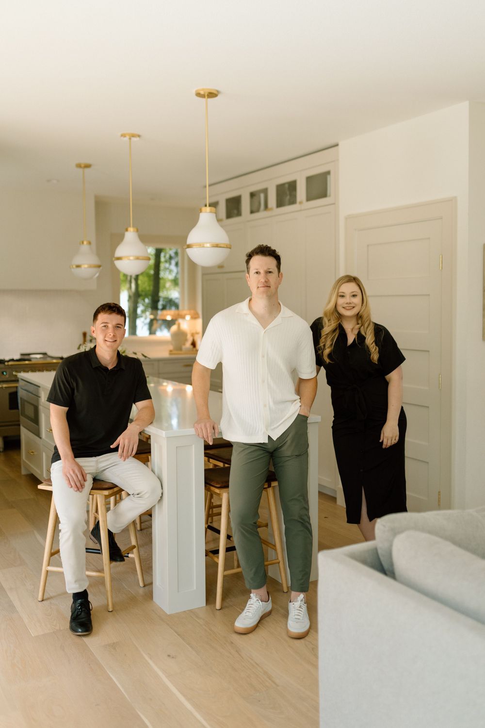 Three people stand casually in modern white kitchen with pendant lights.