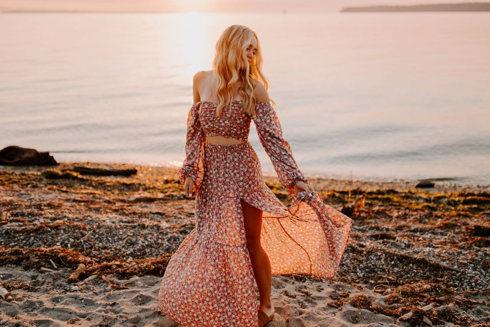 Woman in floral dress walking along rocky beach at sunset carrying woven bag.