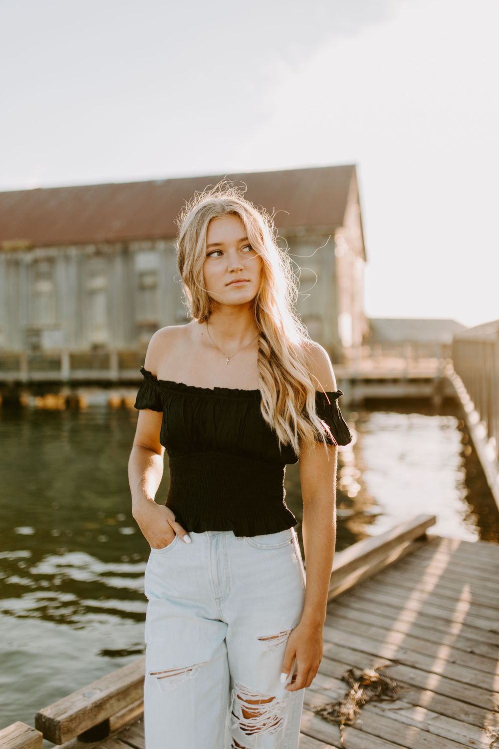 Person in black top and ripped jeans poses on a dock with industrial buildings in background.