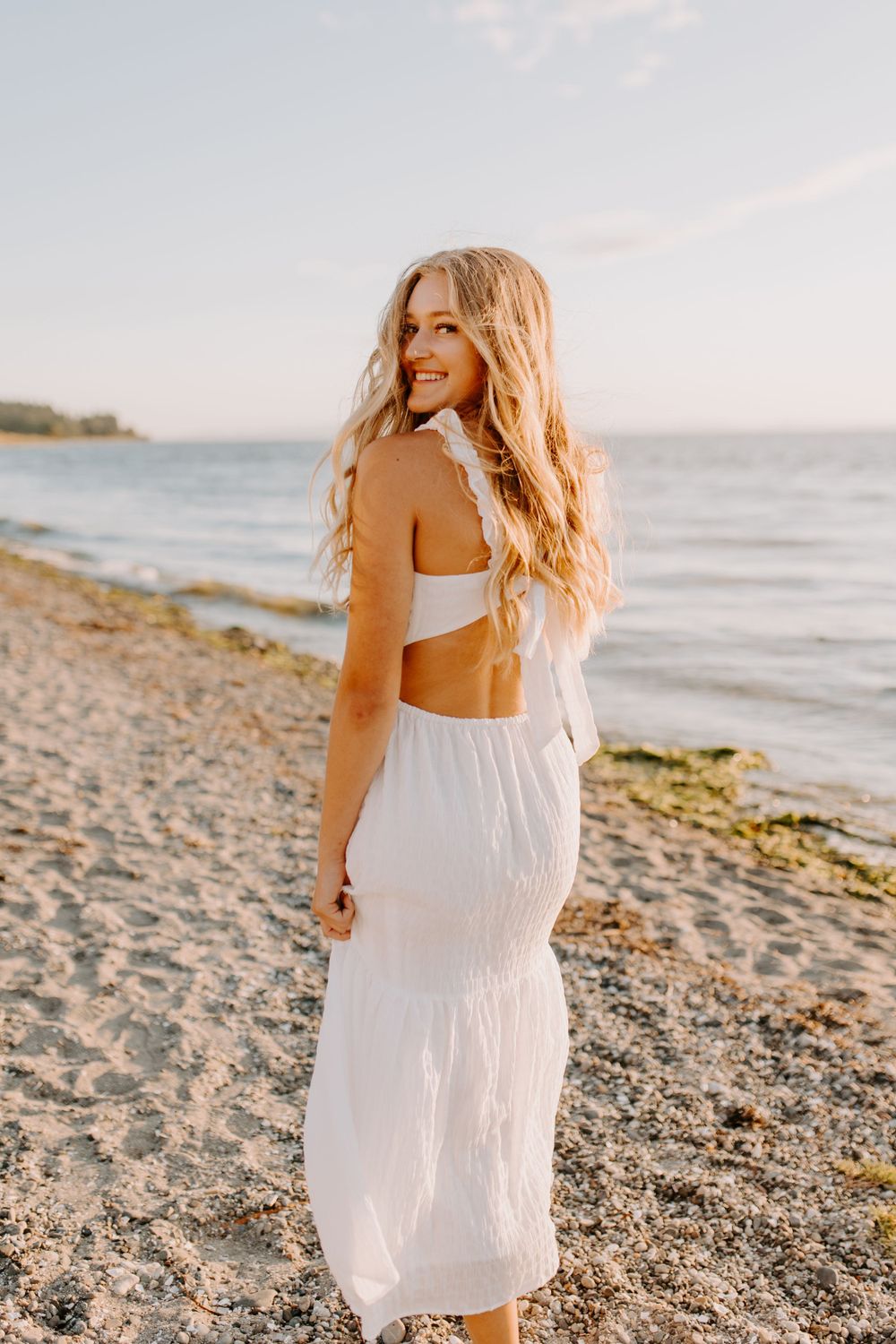 Person in white summer dress poses on beach at sunset with flowing hair.