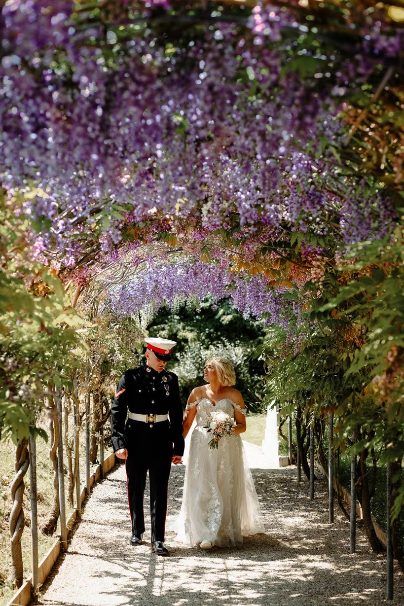 Military Wedding at Deer Park Country House near Honiton. Bride & Groom walking under flower arch