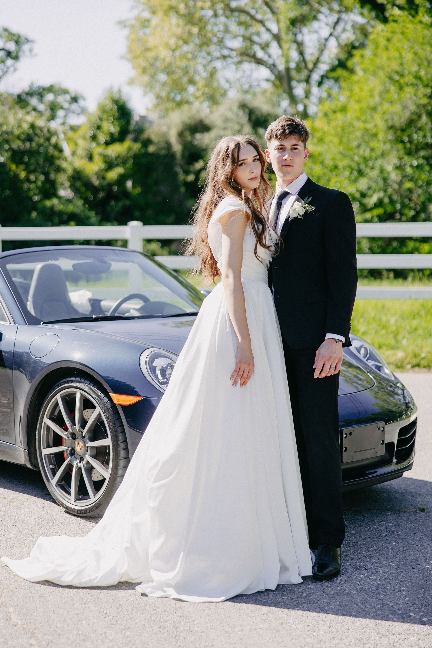 Elegant wedding couple poses beside a navy Porsche sports car outdoors.