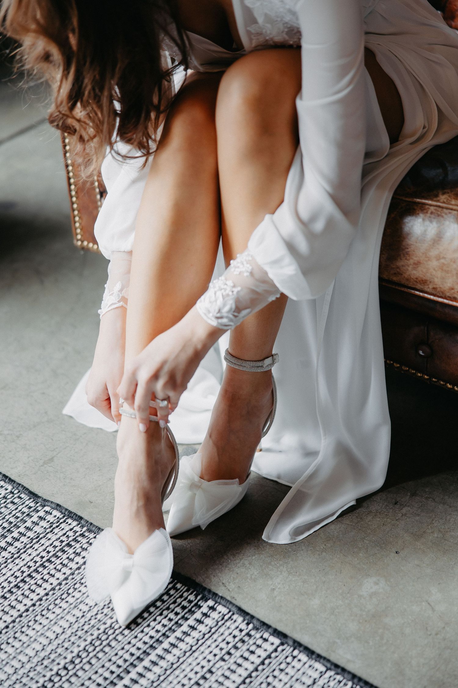 Close-up of white bridal shoes and lace robe detail on leather couch.