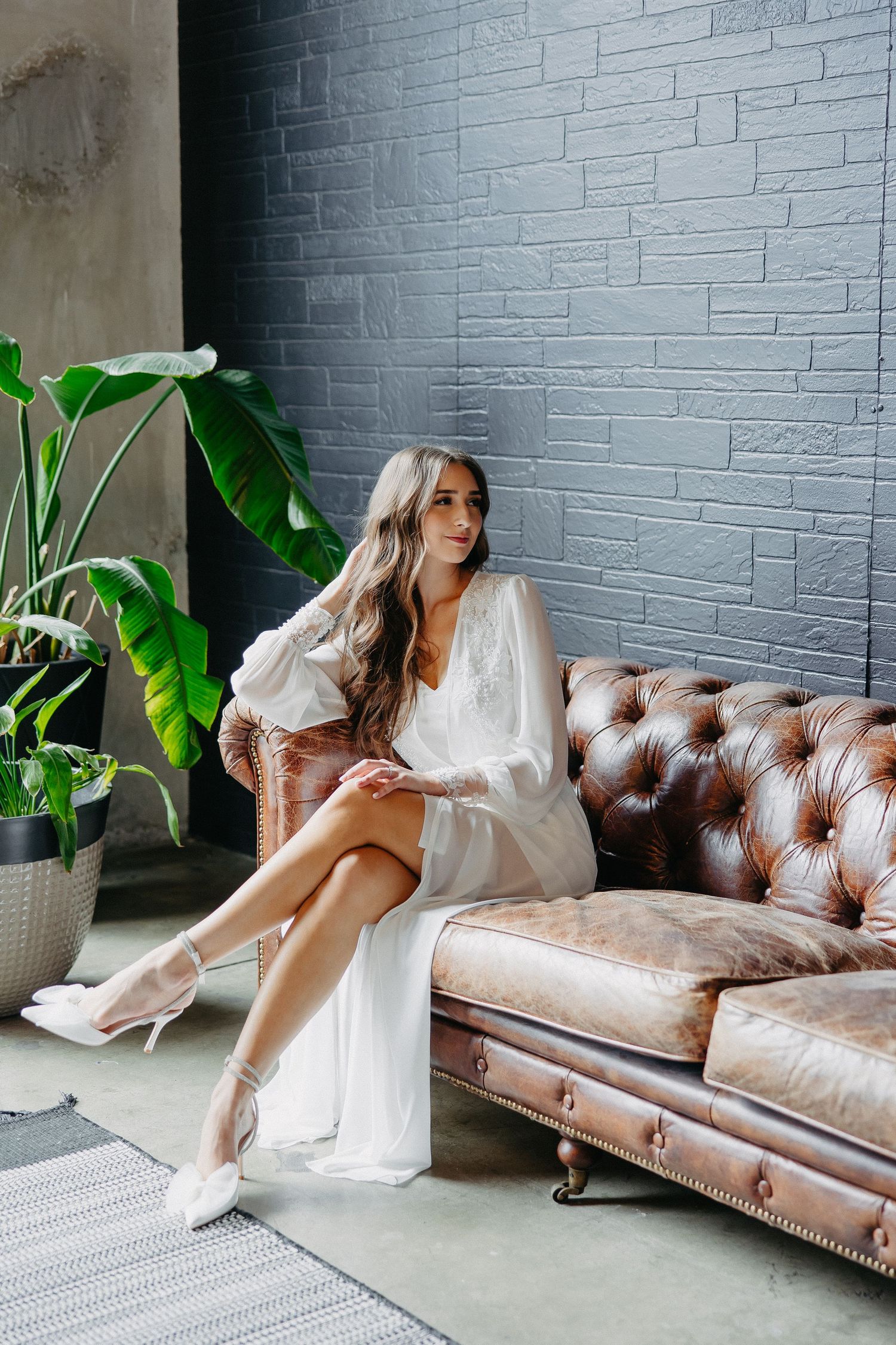 White-robed figure poses elegantly on brown leather couch against industrial brick wall.