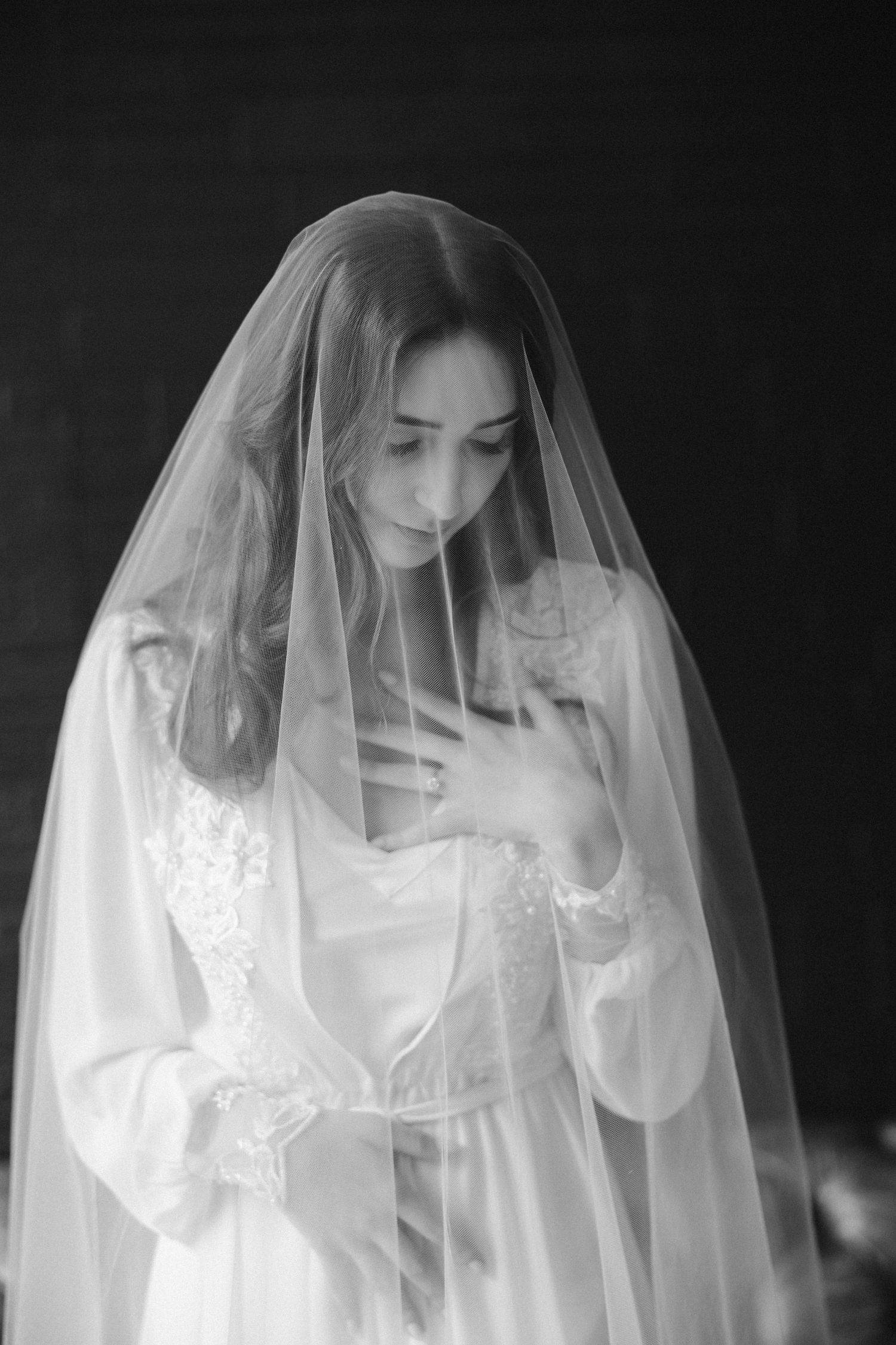 Ethereal black and white portrait of bride in delicate lace veil and robe against dark background.