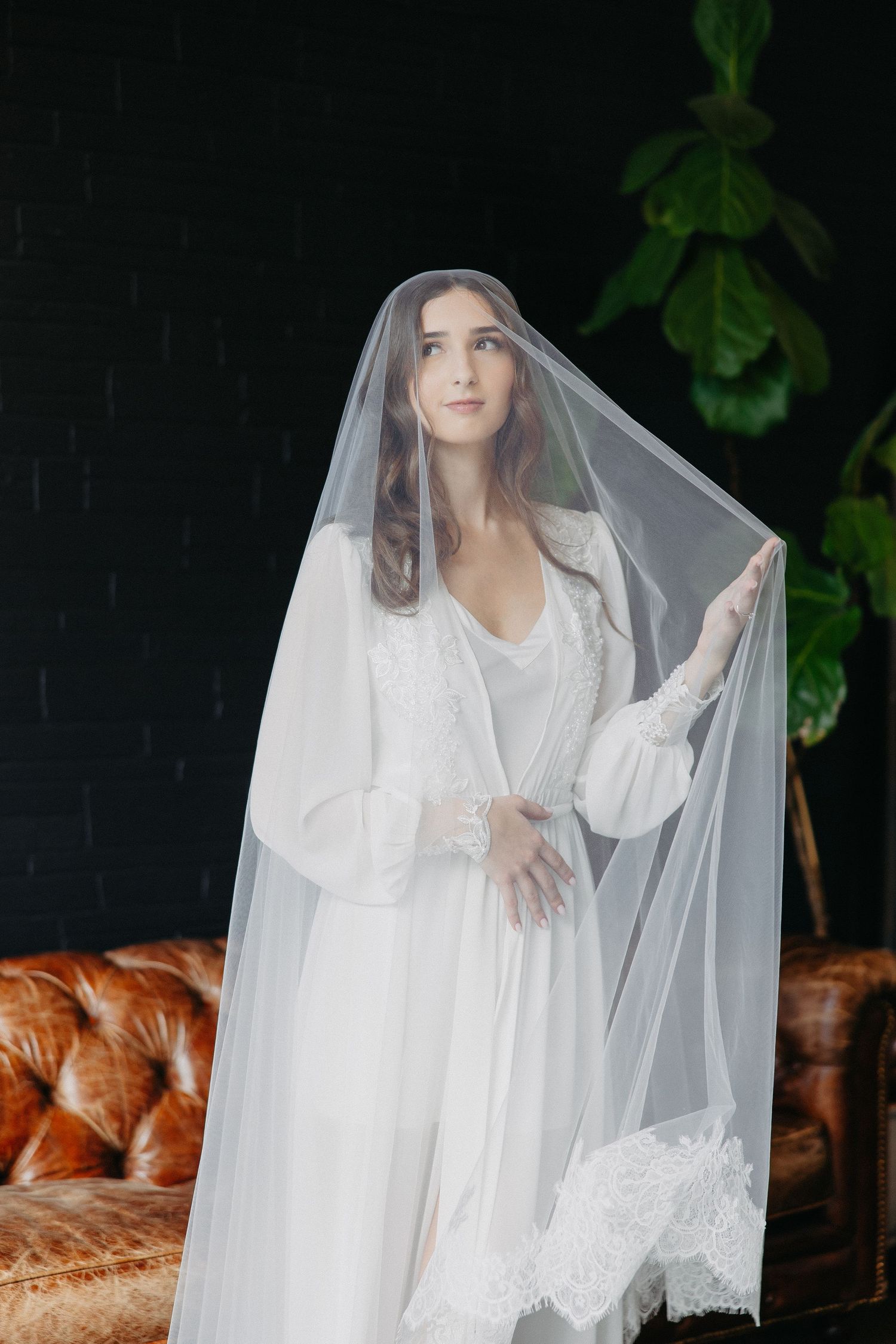 Bride in white robe poses with sheer veil against dark wall with leather couch and green plant.