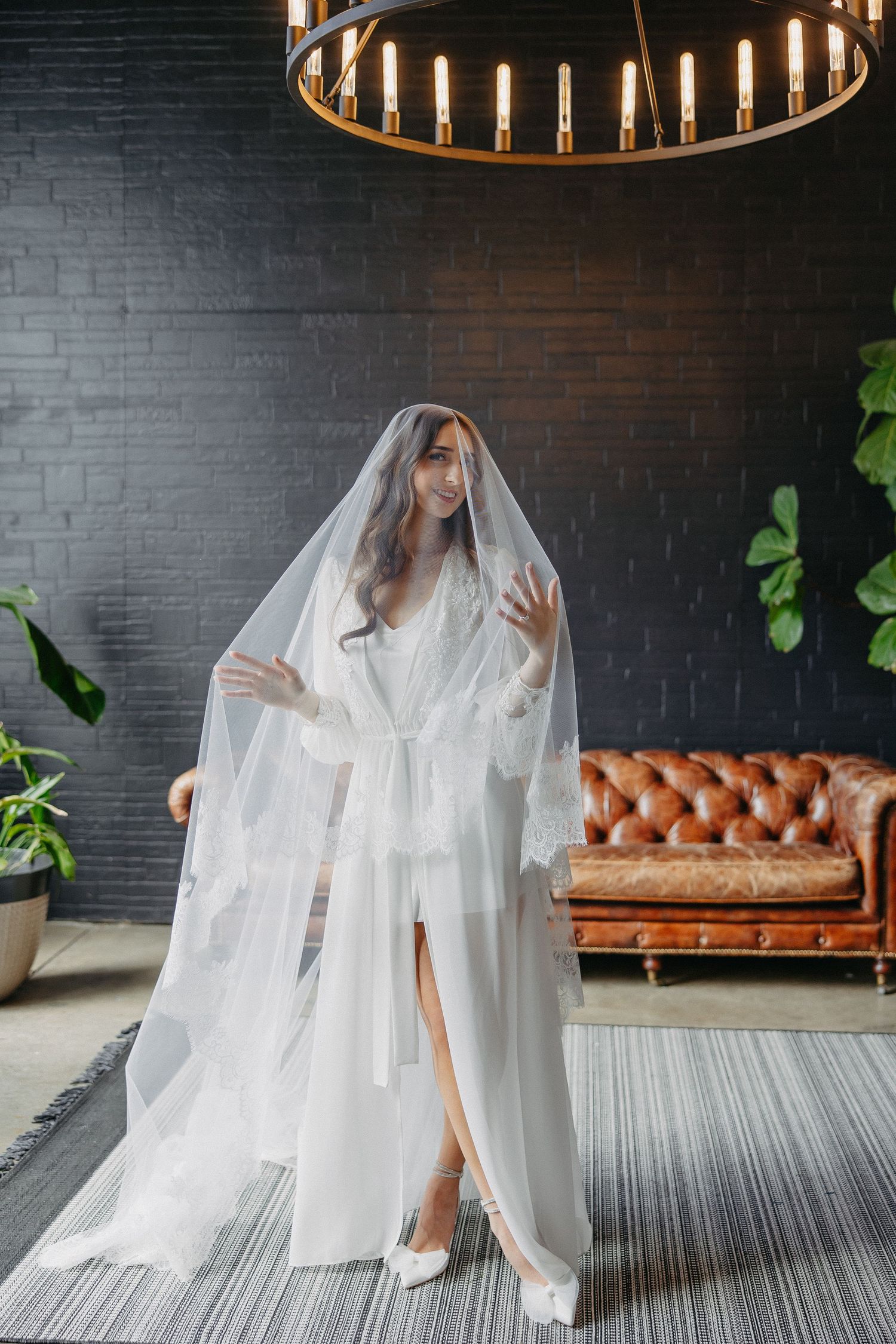 Elegant bridal portrait under circular chandelier with leather couch and brick wall backdrop.