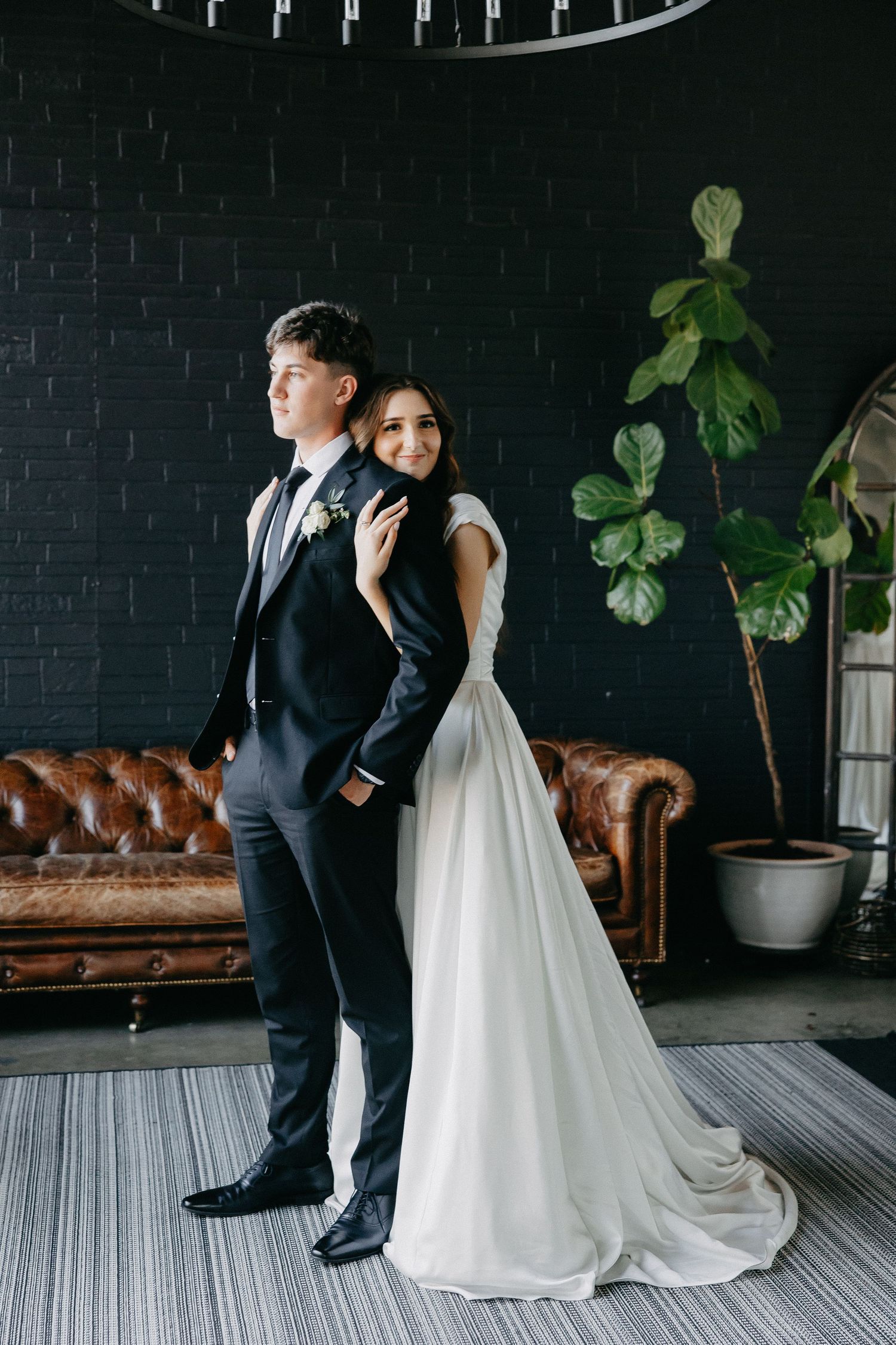 Wedding couple poses against black brick wall with brown leather couch and fiddle leaf fig plant.