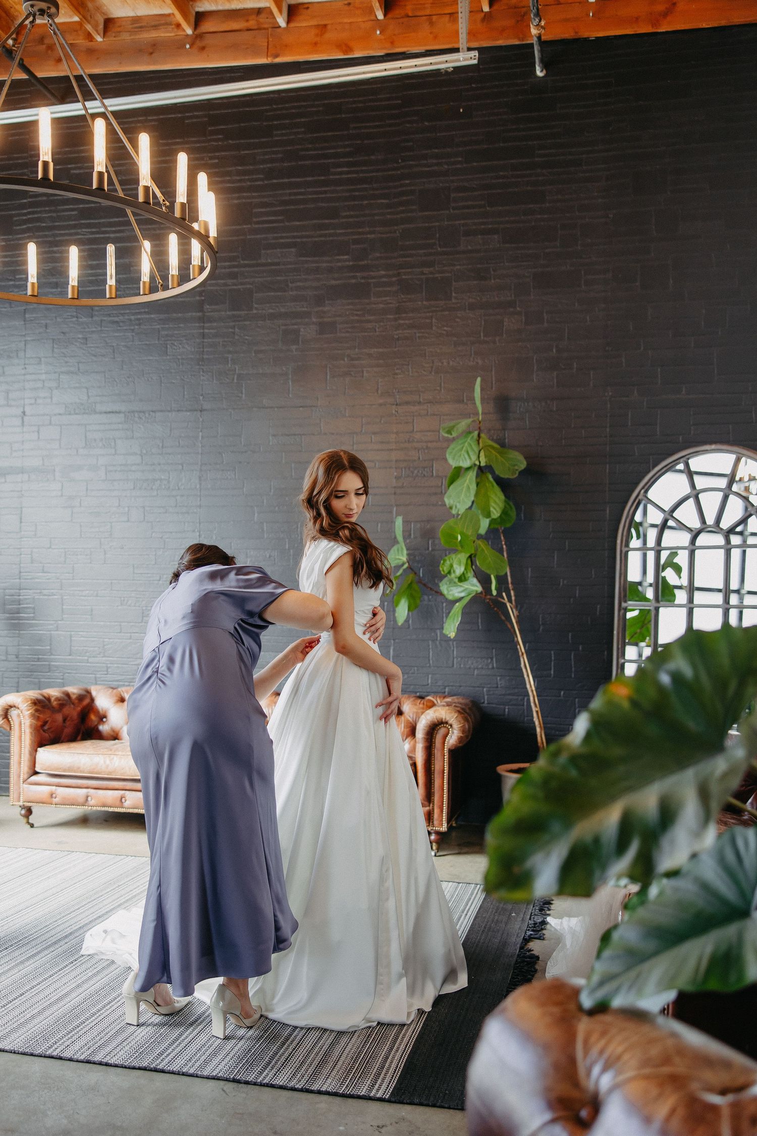 Behind-the-scenes moment of wedding dress fitting in industrial-style room with chandelier.