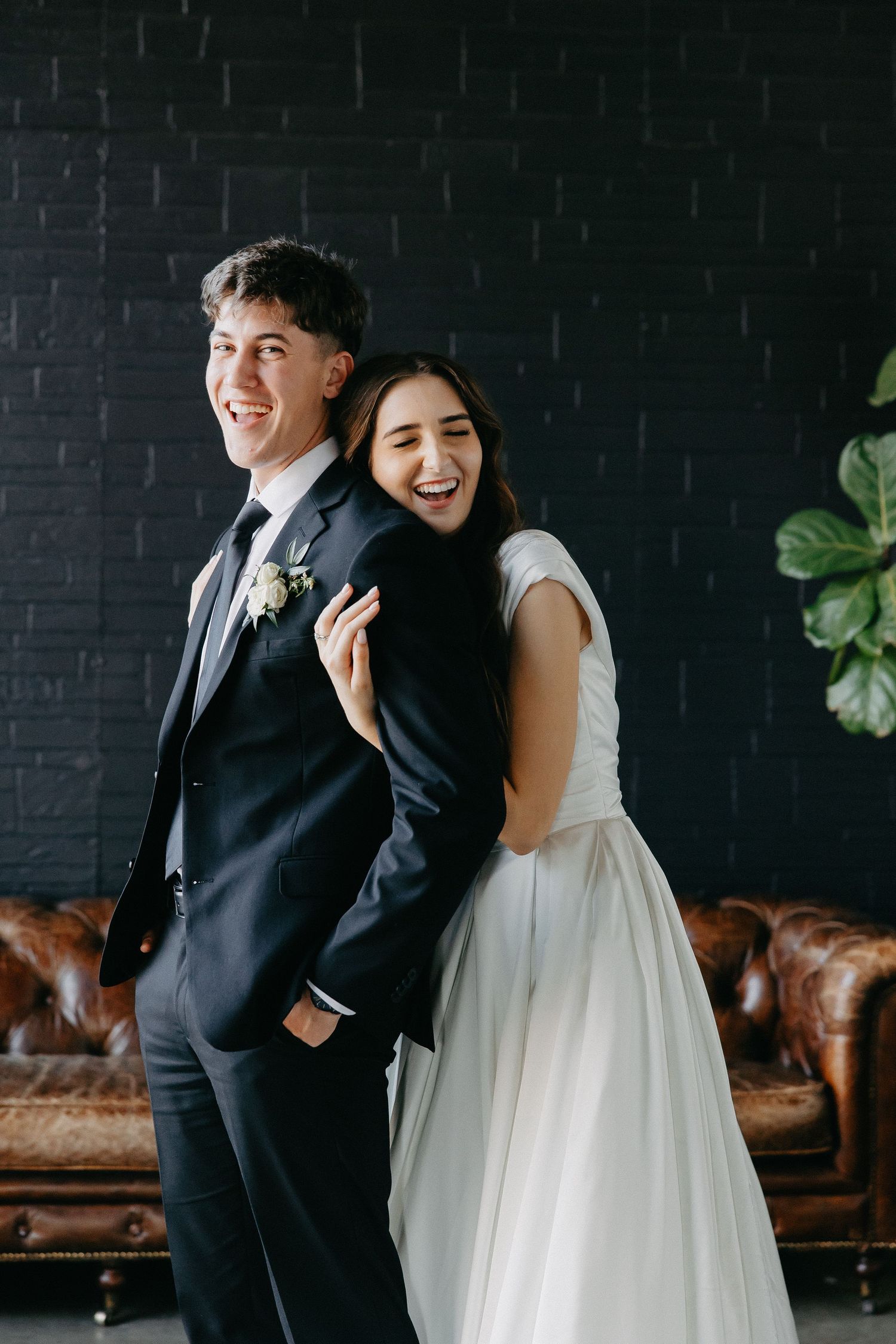 Joyful wedding couple sharing laugh against dark brick wall with vintage leather couch.