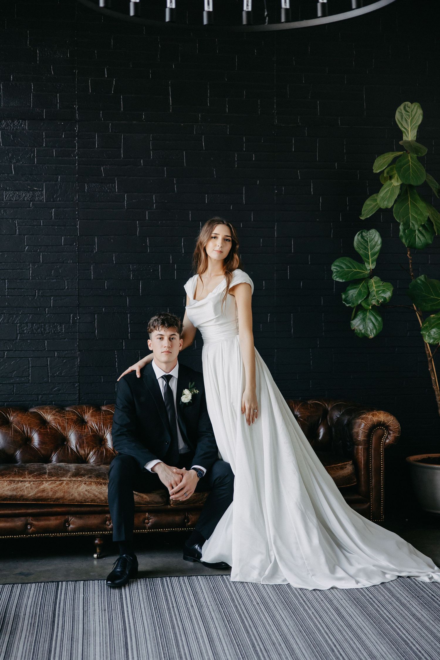 Elegant wedding portrait with couple posed by leather couch against dark brick wall.