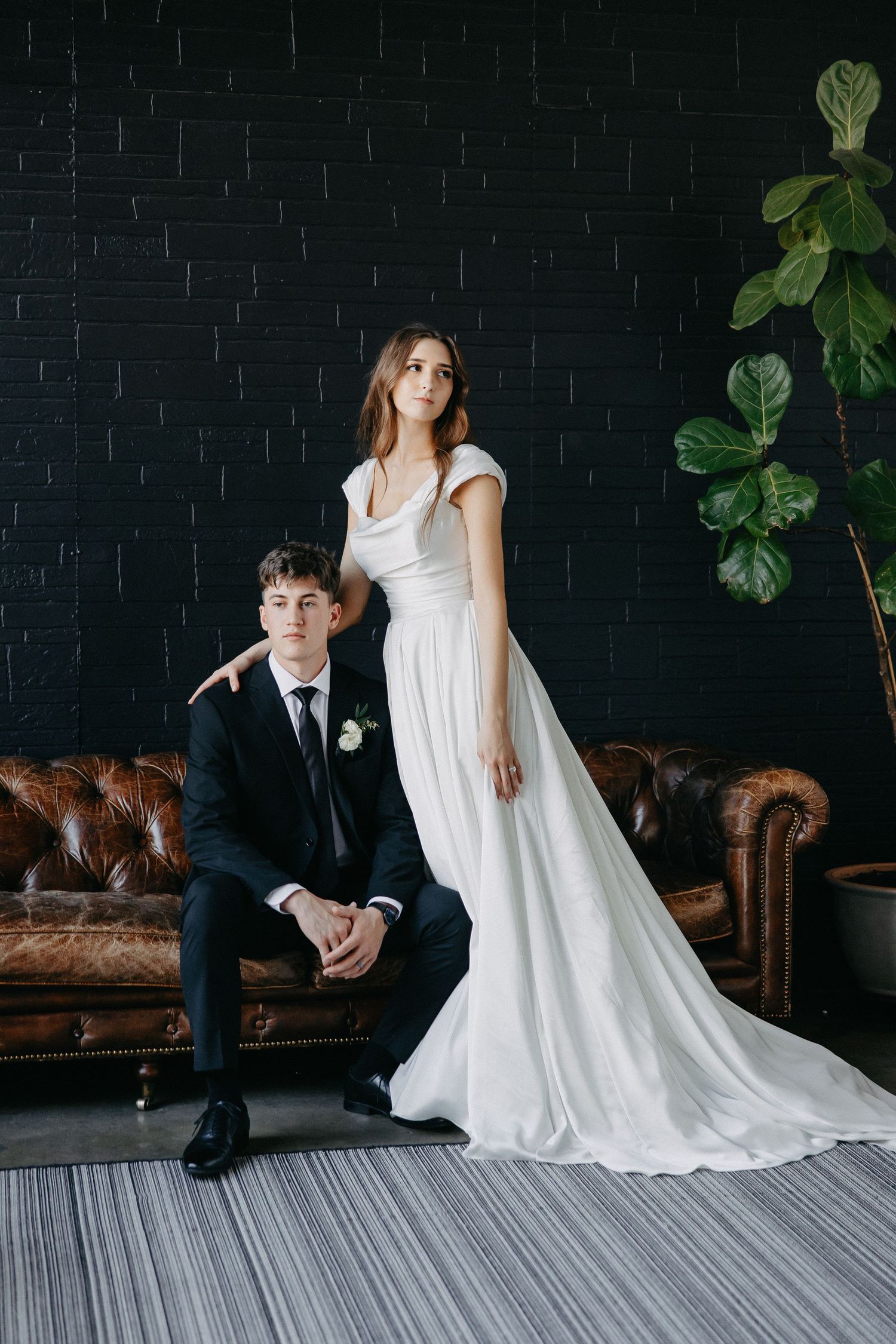 A wedding couple poses against a black brick wall with leather sofas and a fiddle leaf fig plant.