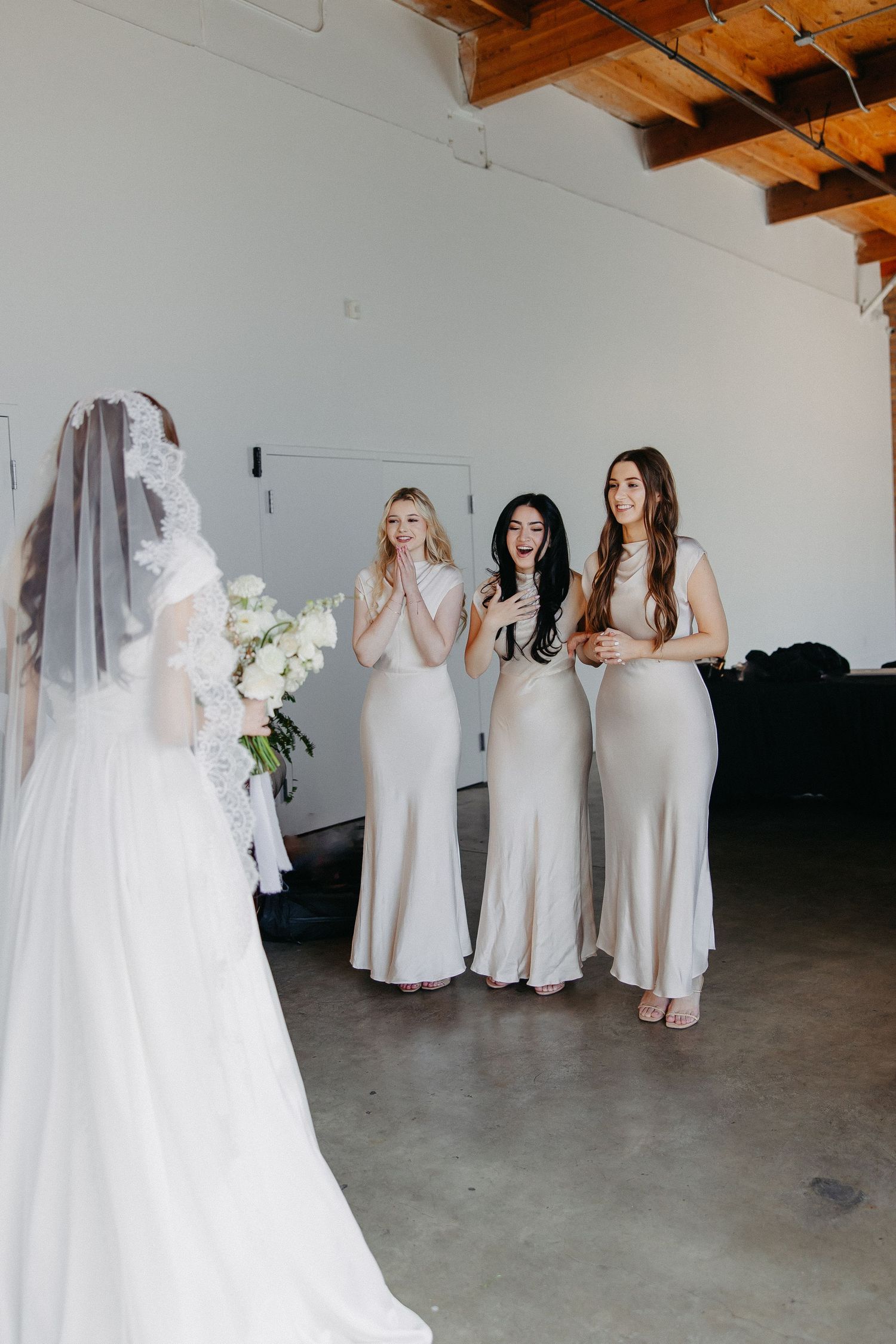 Bridesmaids in cream dresses react emotionally to seeing a wedding gown.