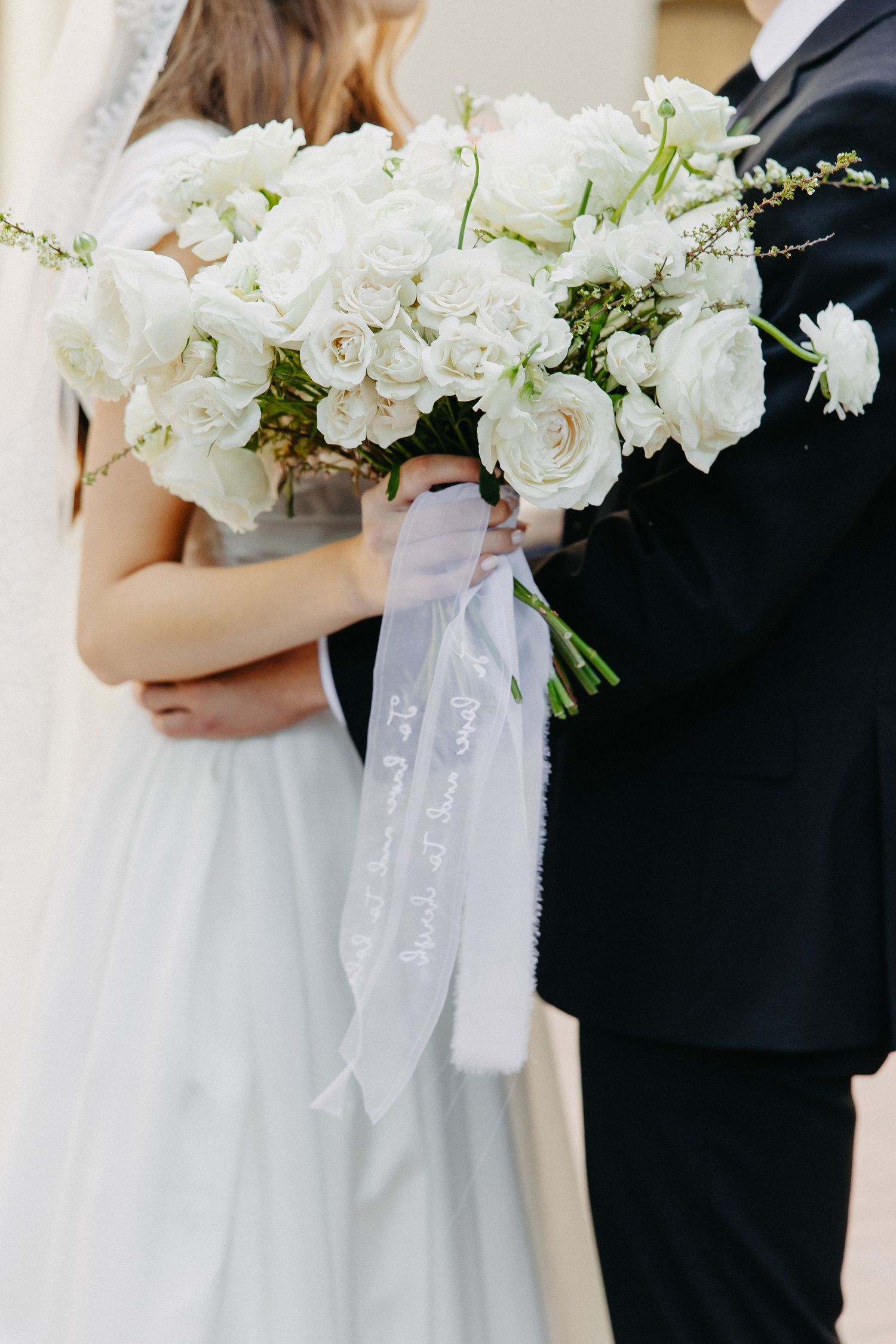 A stunning white rose wedding bouquet with flowing ribbons held between a couple.