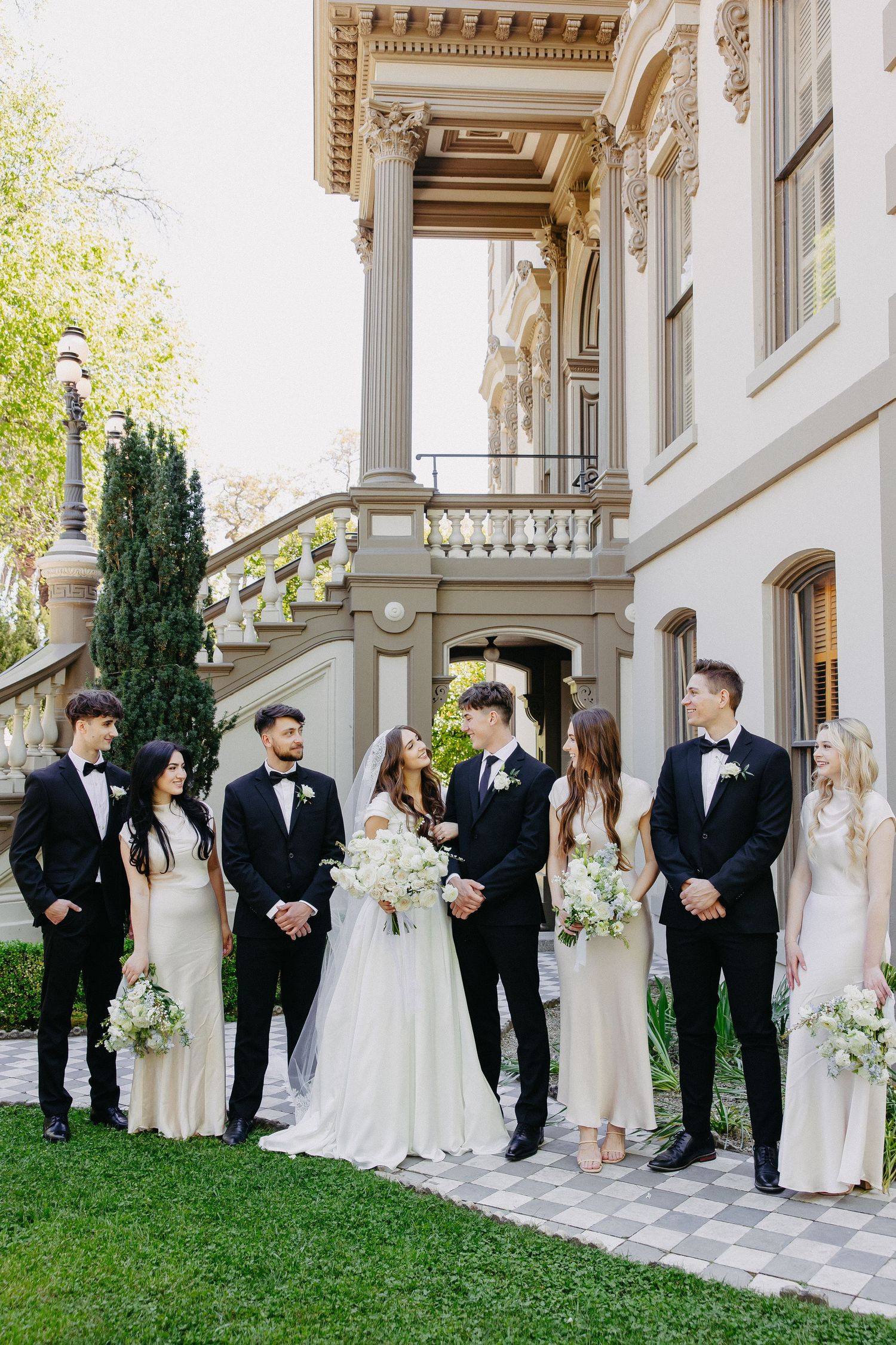 A wedding party poses in formal attire on the steps of a classical building with columns.