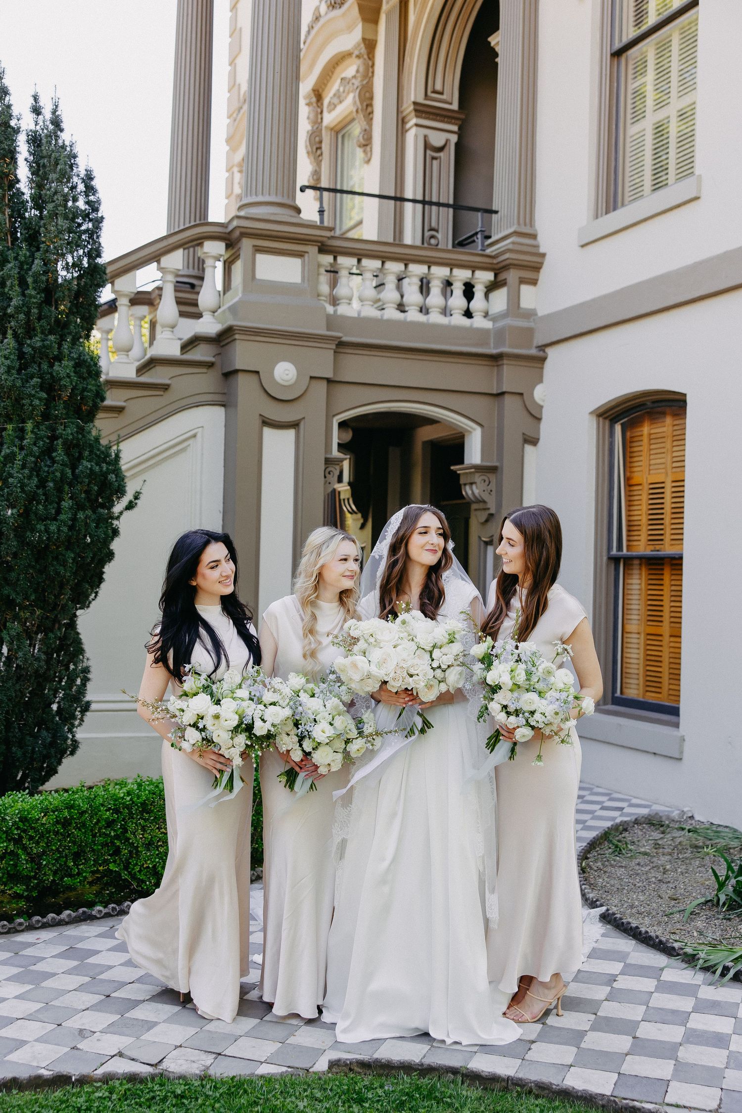 Bridal party with white bouquets poses outside a grand classical building.